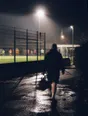 Motion photograph: a man walking away from a floodlit five-a-side pitch at night with his bag, the game still running behind him