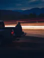 Motion photograph: a man sitting on the open tailgate of a parked pickup at dusk, completely still, traffic running past him