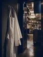 Dark cinematic photograph: Clean chef's whites and a pressed apron hanging ready on a hook in a warm-lit service corridor, the kitchen doors open beyond.