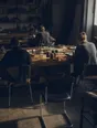 Dark cinematic photograph: An empty chair pulled up to a big table where exactly three others are already working, bright side light.