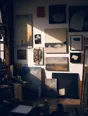 Dark cinematic photograph: A studio wall hung with years of finished work, hard raking light across it.