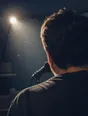 Motion photograph: a man singing at a mic in a small dim rehearsal room, dust moving in the one light