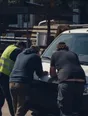 Dark cinematic photograph: Exactly three people leaning over a plan spread on a van bonnet in bright daylight.