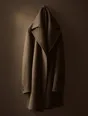 Struck-light photograph: one heavy overcoat hung on a single nail on a bare wall, low light across the nap of the wool