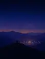Grainy gradient illustration: The lights of a small village clustered close in the valley below, seen from a dark quiet hilltop at nightfall.
