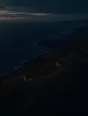Dawn aerial photograph: two stone cottages close together on a dark headland before sunrise, both windows lit.