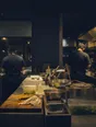 Dark cinematic photograph: Exactly two people working either side of one long prep counter, comfortable distance between them, warm light.