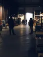 Dark cinematic photograph: Exactly two people talking across a wide warehouse floor while others work on beyond them.