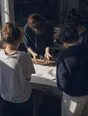 Dark cinematic photograph: Exactly four people standing around a table watching one person's hands move over a drawing.