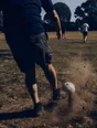 Motion photograph: a man putting the first kick into a park game at midday, the ball just off his foot and dust lifting off the grass
