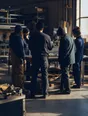 Dark cinematic photograph: Exactly five adults standing in a loose cluster around a workbench in a bright workshop, mid-discussion, the nearest with their back to us and the others listening.