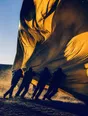 Motion photograph: four men leaning back hard against a huge canvas sheet that is trying to take off in high wind, dust blowing through the light