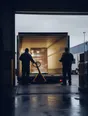 Dark cinematic photograph: Exactly two figures facing each other across a loading bay in flat daylight, standing level.