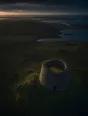 Dawn aerial photograph: a tall roofless round stone tower on a green rise, its grass floor glowing with first light.
