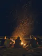 Motion photograph: six men silhouetted around a fire on a dark beach, sparks lifting hard off it into black air