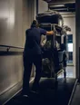 Dark cinematic photograph: A fully loaded trolley being steadied at the top of a ramp, one person's back braced against it.