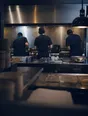 Dark cinematic photograph: A kitchen in the middle of the rush seen from the one still corner of the pass, three cooks moving beyond.