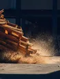 Motion photograph: a stack of timber released onto a yard floor at midday, dust blasting up and lit hard from behind