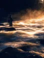 Motion photograph: a man standing still and thigh-deep in a fast shallow river at dawn while the water runs past his legs