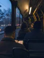 Dark cinematic photograph: A bus interior at dusk, rows of passengers' backs, warm light inside and blue outside.