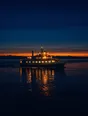 Film photograph: a small lit ferry crossing a calm dark strait at night, its warm lights steady in the water.