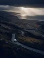Dark cinematic photograph: A hill road switchbacking up out of a valley with one small van climbing it under storm light.