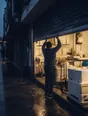 Dark cinematic photograph: A shutter coming down over a bright shopfront at dusk, one person's back beneath it.