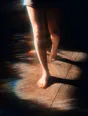 Prism-light photograph: Bare legs walking through hard low sun, the skin and the floor's grain both lit.