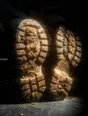 Prism-light photograph: The soles of two boots caked in dried mud, propped up at the end of a walk in hard sun.