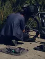 Dark cinematic photograph: A bike chain being re-seated at the roadside in hard sun, the rider crouched with their back to us.