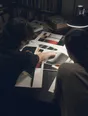 Dark cinematic photograph: Exactly two people leaning over a spread of printed sheets on one working table, one pointing at the page.