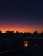 Film photograph: the last stars fading as dawn breaks gold over rooftops, one small kitchen window already lit warm.