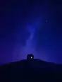 Grainy gradient illustration: One small warm window in a dark house on a hillside, an enormous deep-blue starry sky taking most of the frame.