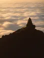 Struck-light photograph: a summit ridge at dawn with a marker cairn on the high point and the world below under cloud