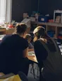 Dark cinematic photograph: Exactly two teachers leaning in over one child's open workbook at a low table, both from behind.