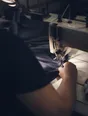 Dark cinematic photograph: A sewing machine mid-seam with the fabric feeding through under the light, one forearm steadying it.
