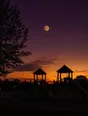 Film photograph: a harvest moon rising over a neighborhood park at dusk, the playground in crisp silhouette below.