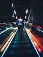 Motion photograph: a man walking fast up a long empty escalator at night, the handrail and ceiling lights running down past him