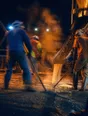 Motion photograph: three men working a concrete pour on a lit site at night, wet concrete running from the chute and steam lifting off the slab