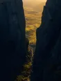 Dawn aerial photograph: a narrow shadowed cleft in dark rock opening onto a vast sunlit valley.