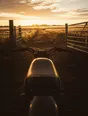 Struck-light photograph: a wide gate standing open onto a field at dawn with a motorcycle waiting, shot from behind it