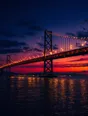 Film photograph: a suspension bridge at dusk, its lights coming on across the whole span above dark reflecting water.