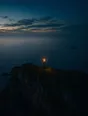 Dawn aerial photograph: a clifftop signal beacon burning small and bright above a dark pre-dawn sea.
