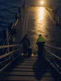 Dark cinematic photograph: A ferry gangway coming down onto a lit quay at dawn, exactly two figures waiting to step off.