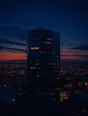 Film photograph: a dark office tower with a few windows still lit, warm home lights glowing across the low city below.
