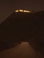 Struck-light photograph: a large modern house lit high on a hill at night, seen from a narrow road a long way below it