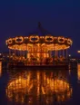 Film photograph: a carousel lit up at blue hour, empty and glowing warm gold, reflected on wet pavement.