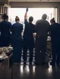 Dark cinematic photograph: One hand raised above a standing group of exactly five at a ward handover, bright morning light.