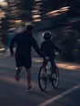 Motion photograph: a man running alongside a child on a bicycle down an empty street at dusk, one hand out behind the saddle
