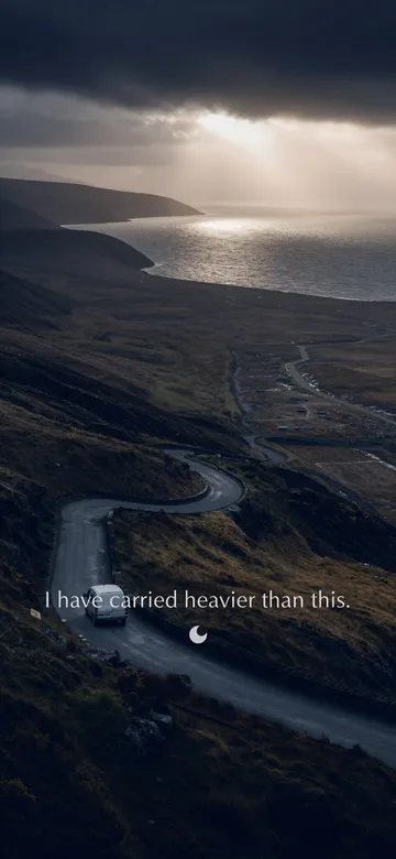 Work affirmations wallpaper: dark cinematic photograph of a hill road switchbacking up out of a valley under storm light, one small van climbing it, with the affirmation 'I have carried heavier than this.' in serif type.